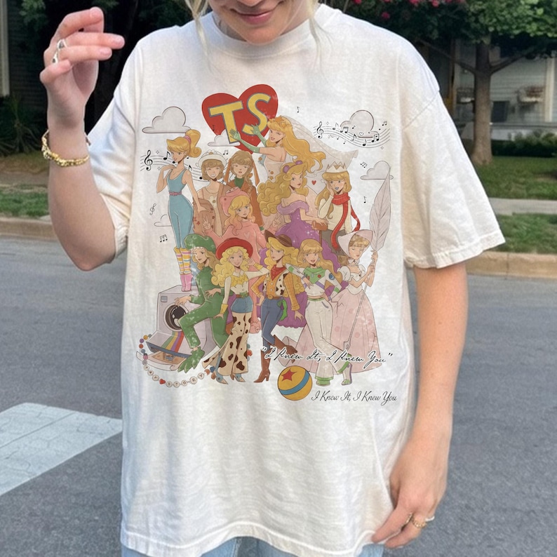 May include: An off-white short-sleeve t-shirt featuring a large graphic print of multiple illustrated female characters, a red heart with the letters TS, a camera, and a ball. The text I Knew It, I Knew You is printed in a cursive script at the bottom.