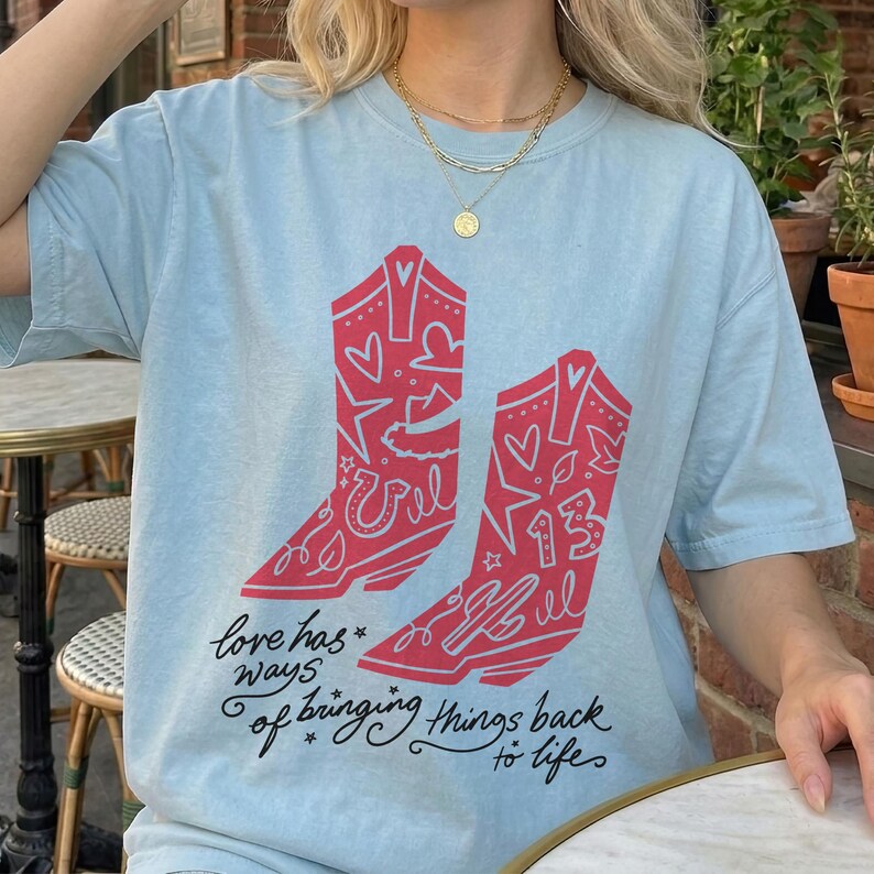K&ouml;nnte beinhalten: Ein hellblaues T-Shirt mit einer Grafik von zwei roten Cowboystiefeln, die mit Herzen, Sternen, einem Hufeisen, einem Kaktus und der Zahl 13 verziert sind. Unter den Stiefeln steht der Text: "love has ways of bringing things back to life."