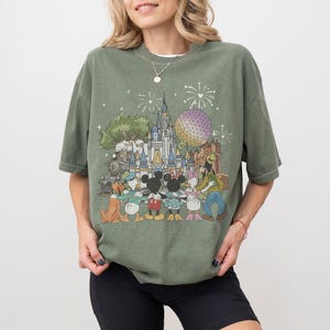 K&ouml;nnte beinhalten: Olivgr&uuml;nes T-Shirt mit einem farbenfrohen Aufdruck eines Vergn&uuml;gungsparks mit Schloss, gro&szlig;er Kugel und Zug. Die Grafik zeigt Zeichentrickfiguren wie Micky und Minnie Maus, Donald Duck und Goofy.