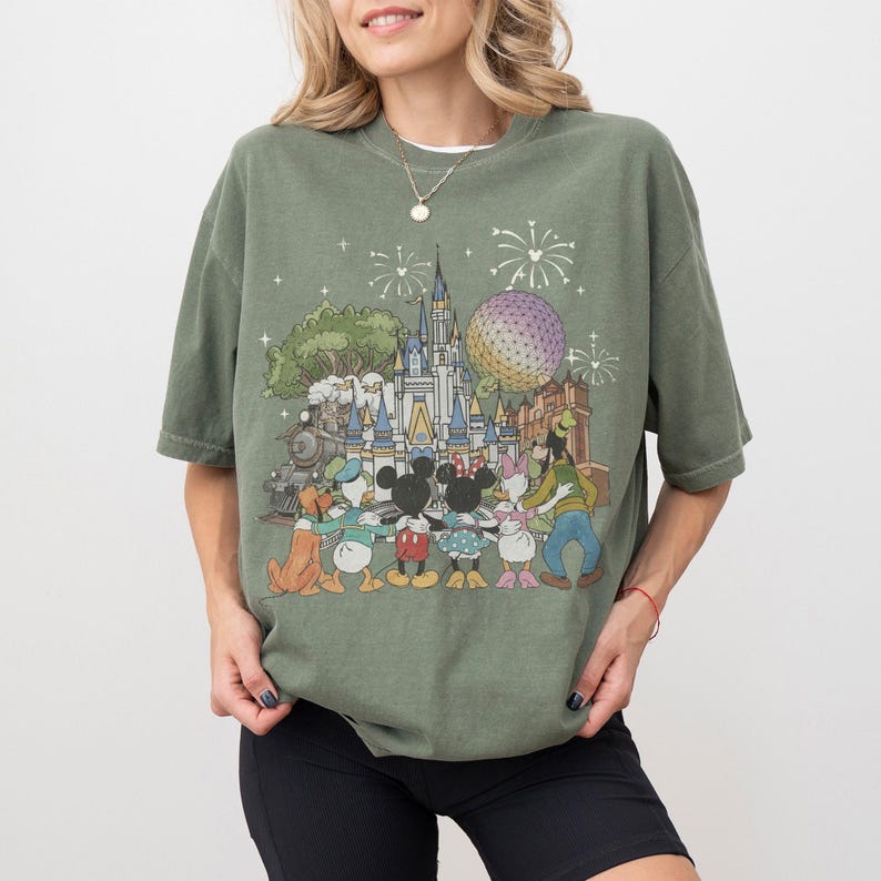 K&ouml;nnte beinhalten: Olivgr&uuml;nes T-Shirt mit einem farbenfrohen Aufdruck eines Vergn&uuml;gungsparks mit Schloss, gro&szlig;er Kugel und Zug. Die Grafik zeigt Zeichentrickfiguren wie Micky und Minnie Maus, Donald Duck und Goofy.