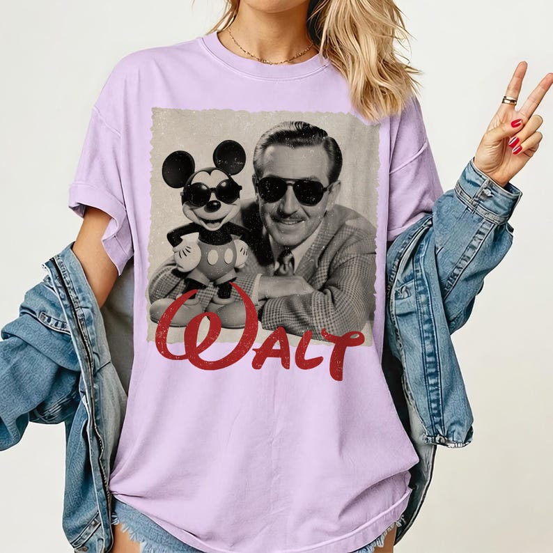 May include: A light purple t-shirt featuring a vintage black and white image of Walt Disney with Mickey Mouse wearing sunglasses. The word "Walt" is printed in red script across the bottom of the image. The shirt has short sleeves.