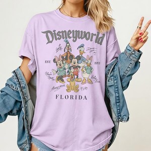 K&ouml;nnte beinhalten: Ein helllila T-Shirt mit dem Text "Disneyworld" und einer Grafik von Disney-Figuren, darunter Micky Maus, Minnie Maus, Donald Duck und Goofy. Der Text "EST. 1971" und "Florida" sind ebenfalls auf dem Shirt aufgedruckt.