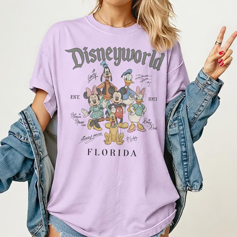 K&ouml;nnte beinhalten: Ein helllila T-Shirt mit dem Text "Disneyworld" und einer Grafik von Disney-Figuren, darunter Micky Maus, Minnie Maus, Donald Duck und Goofy. Der Text "EST. 1971" und "Florida" sind ebenfalls auf dem Shirt aufgedruckt.