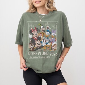 May include: Olive green t-shirt featuring a graphic of Disney characters including Mickey Mouse, Minnie Mouse, Donald Duck, and Goofy. The graphic is framed with the text "Disneyland 2026" and "The Happiest Place on Earth."