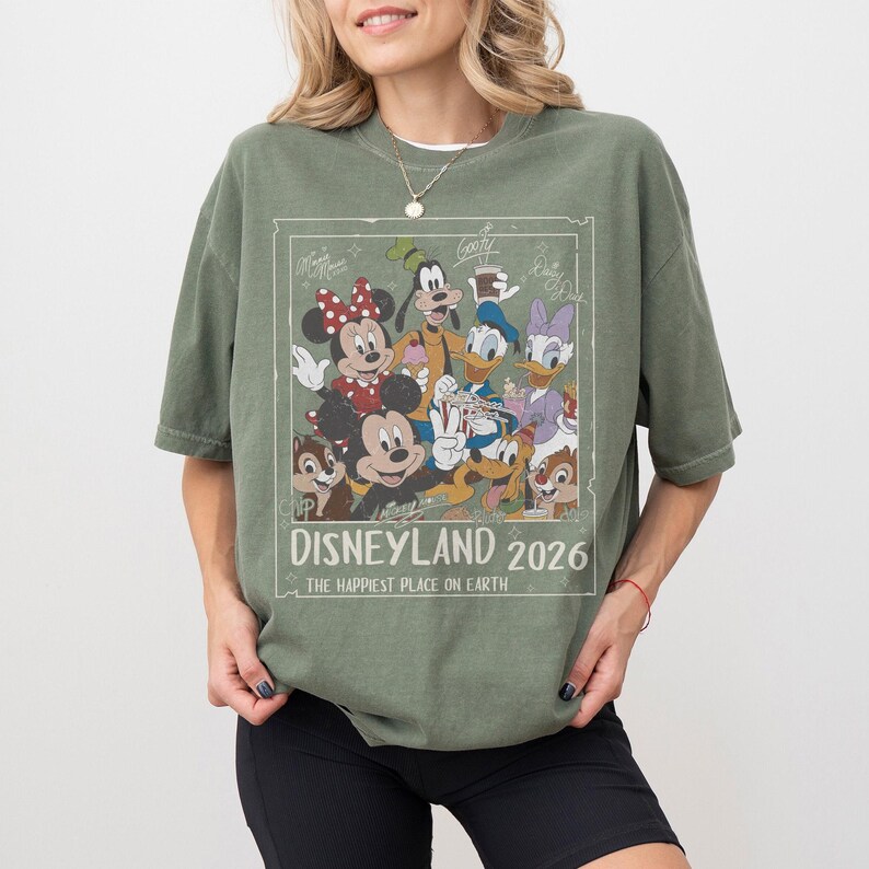 May include: Olive green t-shirt featuring a graphic of Disney characters including Mickey Mouse, Minnie Mouse, Donald Duck, and Goofy. The graphic is framed with the text "Disneyland 2026" and "The Happiest Place on Earth."