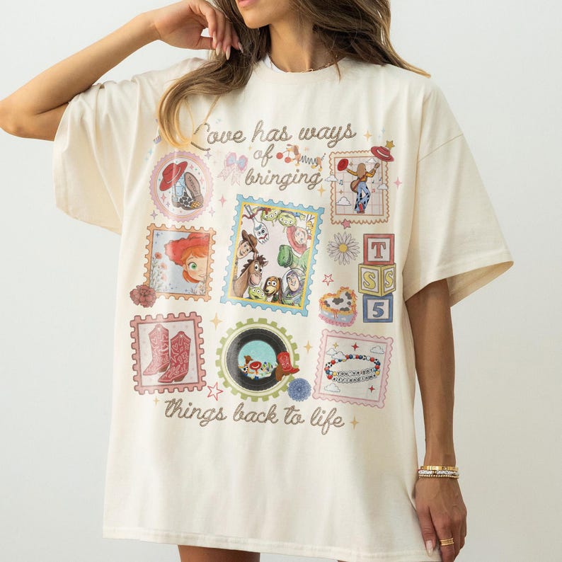 May include: An off-white short-sleeve t-shirt featuring a collage of colorful Toy Story character graphics, postage stamp-style frames, and the text "Love has ways of bringing things back to life" printed in a brown cursive script across the front.