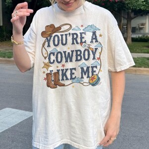 Puede incluir: Una camiseta de manga corta color hueso con un estampado gr&aacute;fico que incluye el texto "YOU'RE A COWBOY LIKE ME" en letras azul oscuro. El dise&ntilde;o presenta un sombrero de vaquero marr&oacute;n, un lazo, una estrella de sheriff, botas, un cohete y un yo-yo azul.