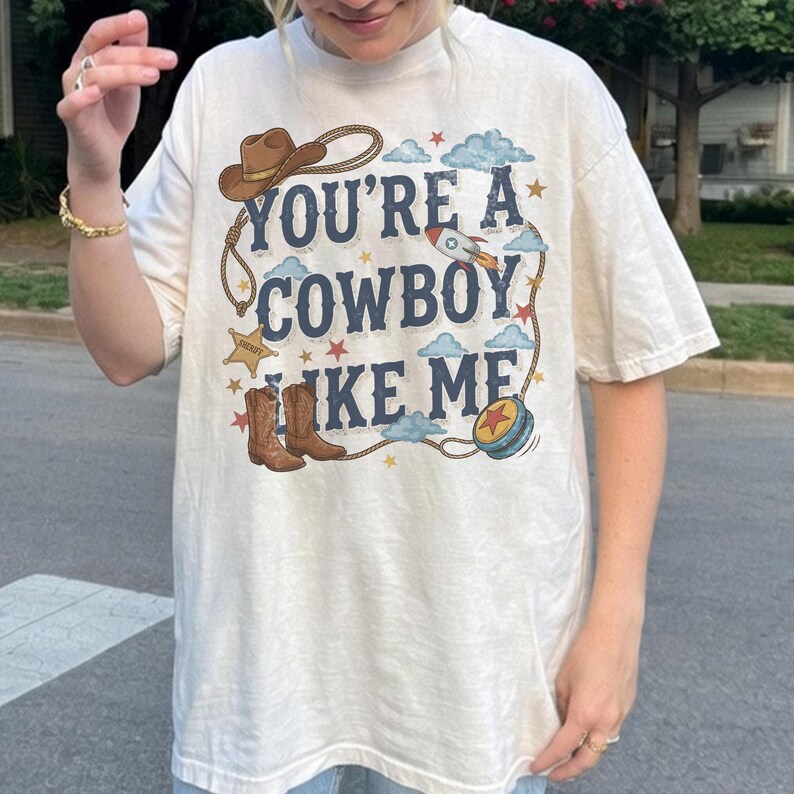 Puede incluir: Una camiseta de manga corta color hueso con un estampado gr&aacute;fico que incluye el texto "YOU'RE A COWBOY LIKE ME" en letras azul oscuro. El dise&ntilde;o presenta un sombrero de vaquero marr&oacute;n, un lazo, una estrella de sheriff, botas, un cohete y un yo-yo azul.