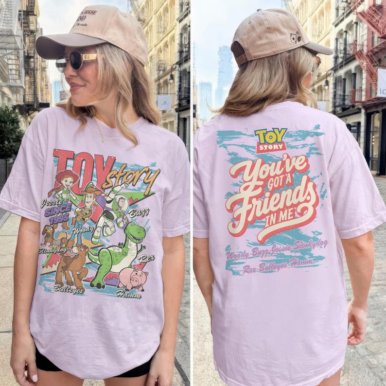 K&ouml;nnte beinhalten: Ein hellviolettes T-Shirt mit Toy Story-Figuren auf der Vorderseite, darunter Woody, Buzz Lightyear, Jessie, Rex, Slinky Dog, Bullseye und Hamm. Die R&uuml;ckseite zeigt den Text "Toy Story You've Got A Friend In Me" mit den Namen der Figuren.