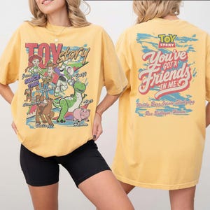 K&ouml;nnte beinhalten: Ein gelbes T-Shirt mit Toy Story-Grafiken. Die Vorderseite zeigt Jessie, Woody, Buzz Lightyear, Slinky Dog, Bullseye und Hamm mit dem Text "TOY STORY SINCE 1995". Die R&uuml;ckseite zeigt den Text "TOY STORY YOU'VE GOT A FRIENDS IN ME".