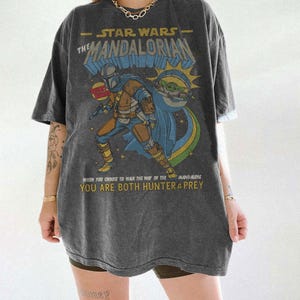 May include: Dark gray t-shirt featuring a graphic of The Mandalorian from Star Wars. The design includes the Mandalorian, Grogu, and text that reads "When you choose to walk the way of the Mandalorian, you are both hunter & prey."
