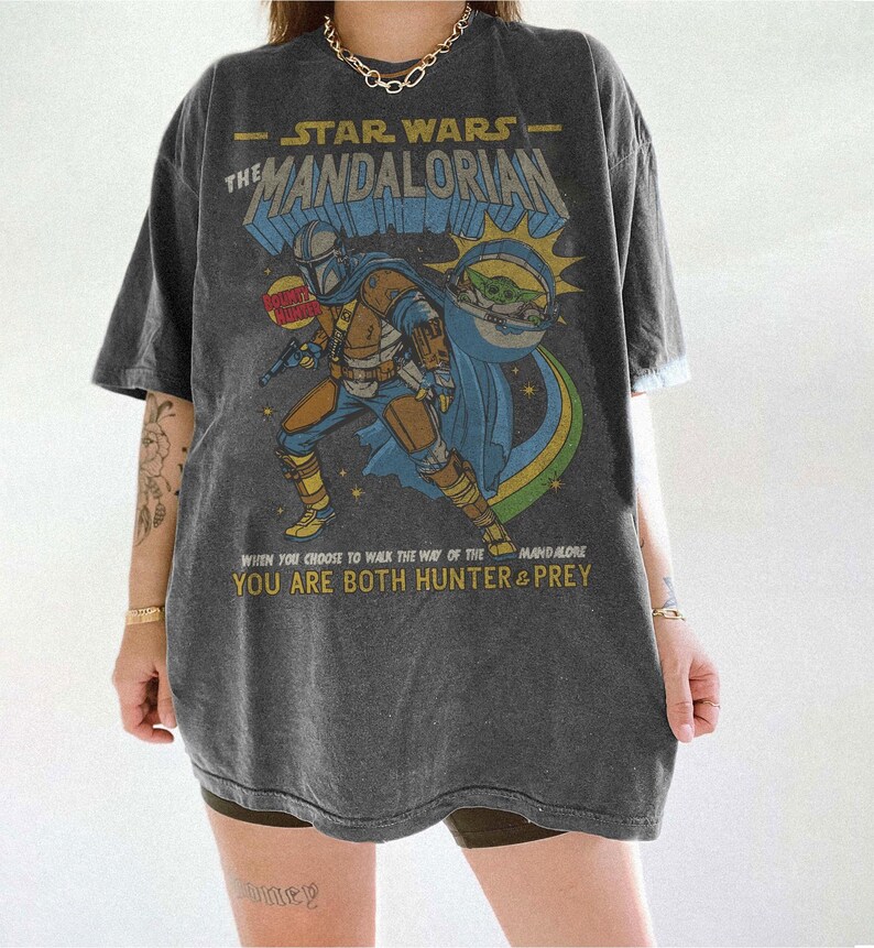 May include: Dark gray t-shirt featuring a graphic of The Mandalorian from Star Wars. The design includes the Mandalorian, Grogu, and text that reads "When you choose to walk the way of the Mandalorian, you are both hunter & prey."