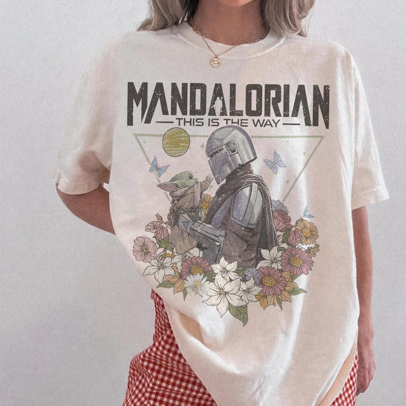 May include: Cream-colored t-shirt featuring a graphic of the Mandalorian and Grogu surrounded by flowers and butterflies. The text "MANDALORIAN - THIS IS THE WAY" is printed across the top in a distressed font.