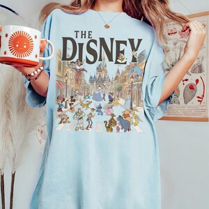 May include: Pale blue t-shirt with the text "THE DISNEY" in brown. The shirt displays a colourful illustration of Disney characters crossing a street, with a castle and buildings in the background.