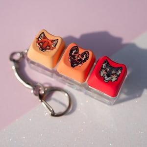 May include: A keychain featuring three square keycaps in a row, colored light orange, medium orange, and red. Each keycap displays a distinct illustrated animal face: a fox, an African wild dog, and a wolf, all rendered in a graphic art style.