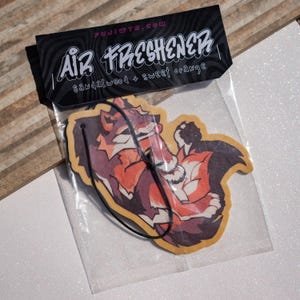 May include: A hanging air freshener in a clear plastic sleeve with a black header card featuring the text AIR FRESHENER, FUJIWTR.COM, and SANDALWOOD + SWEET ORANGE. The freshener is shaped like a stylized orange and dark brown fox with a black string.