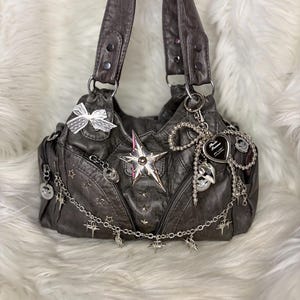 May include: Dark gray handbag with silver star and chain accents. Features a large star, a white lace bow, and a heart-shaped charm. The bag has two handles and a zipper closure.