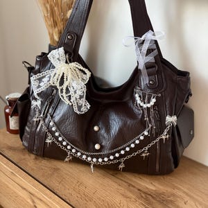 May include: A dark brown leather handbag with multiple pockets and zippers. The bag is adorned with white lace, pearl beads, and silver-colored charms. White ribbons are tied to the straps. The bag is sitting on a wooden surface.