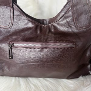 May include: A dark burgundy leather handbag with a zipper pocket and two shoulder straps. The bag has a textured surface and a silver zipper. The background is a fluffy white material.