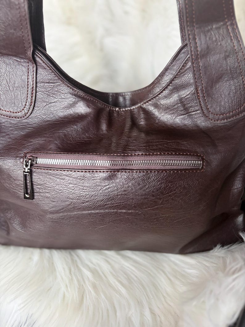 May include: A dark burgundy leather handbag with a zipper pocket and two shoulder straps. The bag has a textured surface and a silver zipper. The background is a fluffy white material.