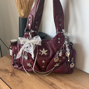 Può includere: Una borsa bordeaux con abbellimenti a stella e ciondoli argentati. La borsa presenta un fiocco, più tasche e un dettaglio a catena. I manici sono attaccati con hardware argentato. La borsa è appoggiata su una superficie di legno.