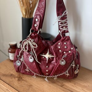 May include: A burgundy handbag with silver embellishments. The bag features a large star, smaller stars, and silver charms. It has a shoulder strap and is decorated with silver studs and a chain.