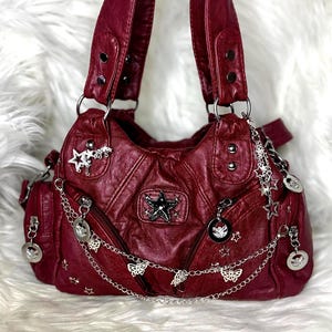 May include: A burgundy faux leather handbag with silver-tone hardware. The bag features multiple pockets, decorative charms, and a chain with butterfly accents. The bag has two shoulder straps.