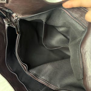 May include: A close-up view of a dark brown leather handbag with a zipper closure. The interior of the bag is visible, revealing a gray fabric lining and several pockets. The bag has a handle and a shoulder strap.