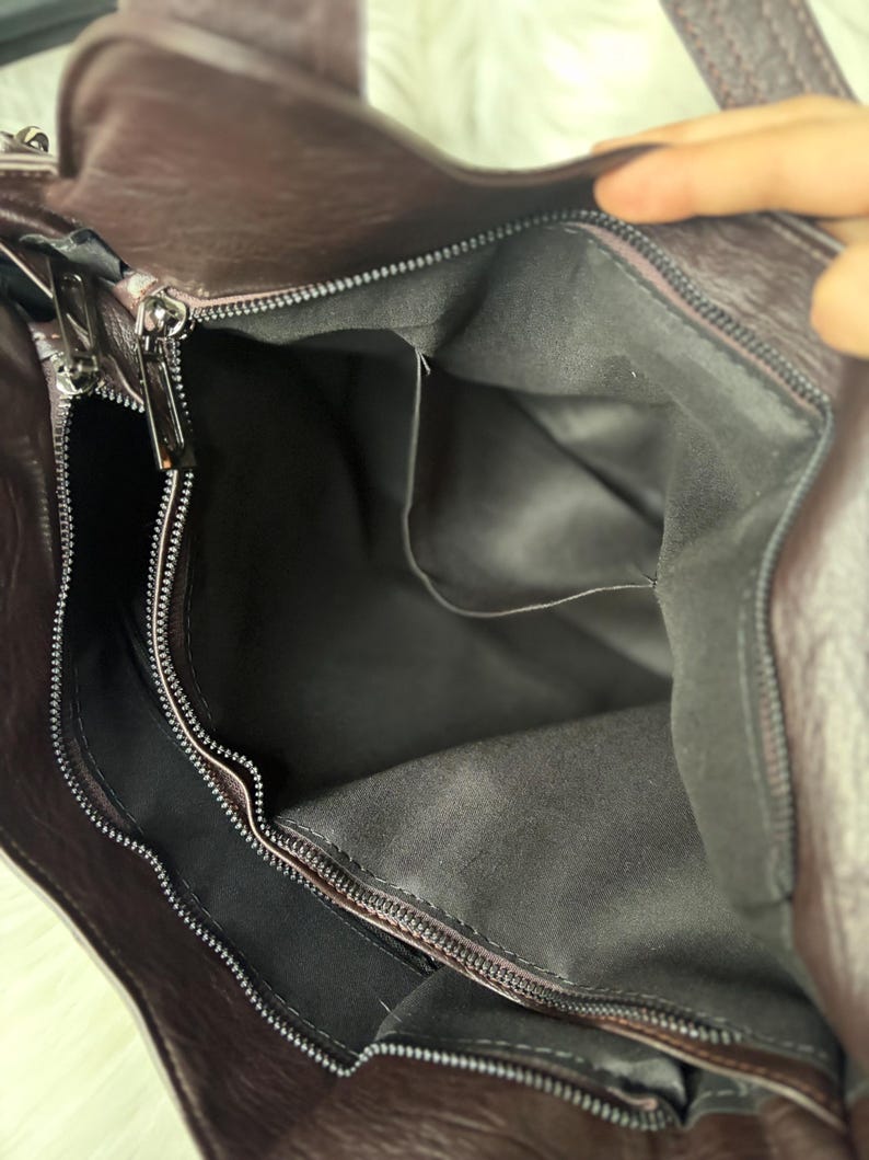 May include: A close-up view of a dark brown leather handbag with a zipper closure. The interior of the bag is visible, revealing a gray fabric lining and several pockets. The bag has a handle and a shoulder strap.