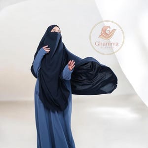 May include: A woman wearing a navy blue niqab and a matching long dress. The niqab covers the face, with a flowing fabric extending from the head covering. The dress is a solid blue color. The logo for Ghanirra Scarves is in the background.