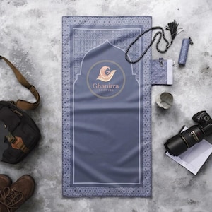 May include: A blue prayer mat with a decorative border and the "Ghanirra Scarves" logo. A black camera, prayer beads, and other accessories are arranged around the mat on a gray surface.