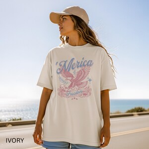 May include: Ivory-colored t-shirt with a graphic design featuring a pink eagle, blue text that says "'Merica", and the word "Freedom" on a banner. The shirt is paired with blue shorts and a tan baseball cap.
