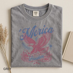 May include: Grey t-shirt with a distressed graphic featuring a pink eagle, blue text that says "'Merica" and "Freedom". The shirt has a crew neck and a tag that says "Comfort Colors".