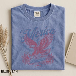 May include: A blue jean colored t-shirt with a crew neck. The shirt features a graphic of an eagle with the word "'Merica" above it and the word "Freedom" below it. The shirt has a vintage, distressed look.