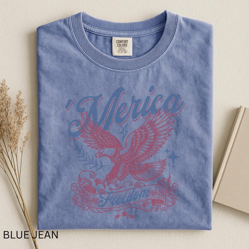 May include: A blue jean colored t-shirt with a crew neck. The shirt features a graphic of an eagle with the word "'Merica" above it and the word "Freedom" below it. The shirt has a vintage, distressed look.