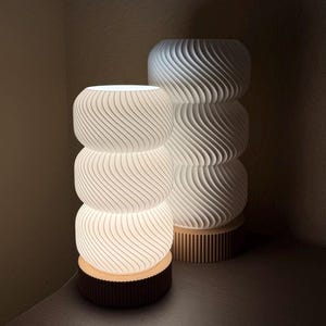 May include: Two white, stacked, spiral-patterned lamps with a warm glow. The lamps are cylindrical, with a series of curved lines creating a textured effect. One lamp sits on a dark brown base, the other on a light brown base. The lamps are on a wooden surface.