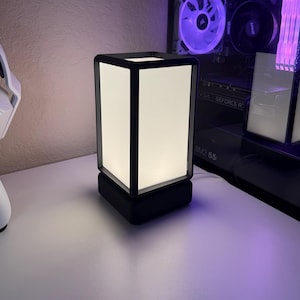May include: A rectangular table lamp featuring a solid black frame and a white translucent shade that emits a soft glow. The lamp has a square black base and a clean, geometric design, providing illumination through its four vertical panels.