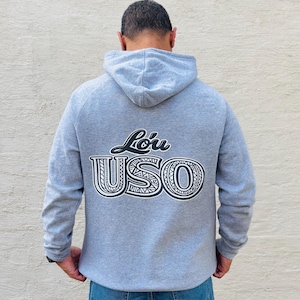 May include: A grey hoodie featuring the text "Lō'u USO" in a black and white design on the back. The hoodie has a hood and long sleeves. The person in the image is wearing blue jeans.