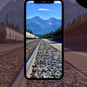 May include: A smartphone displays a scenic train track view with mountains and a blue sky. The image shows a train station with a train on the left and a platform on the right. The phone has a black border and the text "ORDER NOW" on a button.
