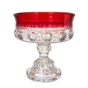 May include: A clear glass pedestal bowl featuring a vibrant red rim and a textured surface with raised circular dot patterns. The bowl stands on a sturdy, matching glass base with a repeating dot design that extends from the stem to the foot.