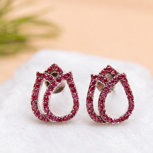 Puede incluir: Un par de pendientes de botón con forma de tulipán, con un marco metálico incrustado con pequeñas piedras de color rosa vibrante. Los pendientes tienen un centro hueco y un cierre de presión, mostrando un diseño floral detallado en rosa.