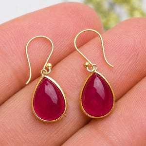 Peut inclure: Une paire de boucles d'oreilles pendantes dorées avec des pierres lisses en forme de goutte d'un rose profond. Chaque boucle est suspendue à un crochet en fil doré incurvé, présentant une monture en métal poli entourant la pierre vive.