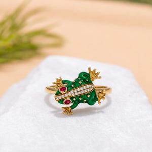 May include: A gold ring featuring a green enameled frog design. The frog has two red circular eyes, a back stripe encrusted with small clear stones, and textured gold feet. The band is a simple gold metal finish, showcasing a detailed animal motif.