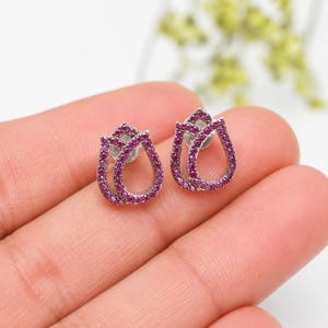 May include: A pair of stud earrings shaped like tulips, featuring a silver-toned metal frame encrusted with small, vibrant pink stones. Each earring has a hollow center and a secure post backing, showcasing a delicate floral silhouette design.