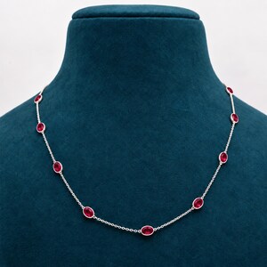 Può includere: Una delicata collana a catena argentata con pietre preziose rosse di forma ovale, distanziate uniformemente e incastonate in castoni di metallo lucido. La collana presenta una serie di queste vivaci pietre color cremisi collegate da sottili maglie metalliche, creando un design raffinato e simmetrico.