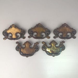 May include: Five brass-toned metal drawer pulls with a decorative, ornate shape and a swinging bail handle. Each piece features a central mounting plate with curved, scroll-like edges and a dark, aged patina finish on the metal surface.