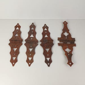 May include: Four antique, rusty metal hinges with decorative cut-out designs. The hinges are a reddish-brown color and appear to be aged. The hinges are likely for a door or cabinet.
