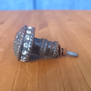 May include: A decorative cabinet knob with a bronze finish and a rope-like texture. The knob is adorned with a ring of clear, sparkling rhinestones. A threaded bolt and nut are attached to the back.