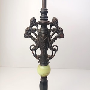 May include: An antique-style lamp finial in dark bronze metal, featuring an elaborate floral design. It has a light green, spherical accent and sits on a dark, rectangular base. The finial's design is detailed and vintage-inspired.