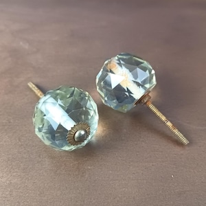 Puede incluir: Dos pomos de armario de cristal transparente facetado con herrajes color latón. Los pomos son redondos y tienen un patrón de diamante. Los tornillos están oxidados y el fondo es de color marrón liso.